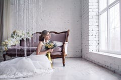 La belle jeune mariée dans la robe de mariage blanche s'assied près du sofa devant la fenêtre et tient le bouquet photo stock La belle jeune mariée dans la robe de mariage blanche s'assied près du sofa devant la fenêtre et tient le bouquet photo stock