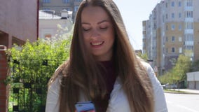 La chica joven hermosa escribe un mensaje para utilizar un smartphone que camina abajo de la calle en el verano y la sonrisa almacen de metraje de vídeo La chica joven hermosa escribe un mensaje para utilizar un smartphone que camina abajo de la calle en el verano y la sonrisa almacen de metraje de vídeo