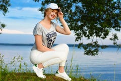 La chica joven romántica en deportes viste con un casquillo en la luz del sol contra el contexto de la orilla del agua del lago e Imagen de archivo La chica joven romántica en deportes viste con un casquillo en la luz del sol contra el contexto de la orilla del agua del lago e Imagen de archivo