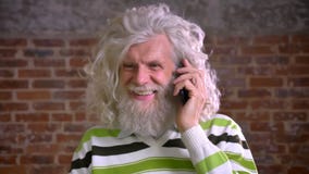 La conversación tranquila sobre el teléfono del viejo varón caucásico con la gran barba blanca y el pelo ondulado, enfocó o su ll almacen de metraje de vídeo La conversación tranquila sobre el teléfono del viejo varón caucásico con la gran barba blanca y el pelo ondulado, enfocó o su ll almacen de metraje de vídeo