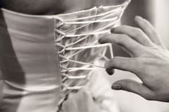 La damigella d'onore sta legando il vestito da sposa, il corsetto della sposa dalla parte posteriore Fotographia in bianco e nero immagini stock La damigella d'onore sta legando il vestito da sposa, il corsetto della sposa dalla parte posteriore Fotographia in bianco e nero immagini stock