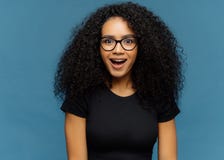 La femme pelée foncée agréablement étonnée maintient la mâchoire lâchée, des regards fixes avec l'intérêt, a les cheveux bouclés, photos stock La femme pelée foncée agréablement étonnée maintient la mâchoire lâchée, des regards fixes avec l'intérêt, a les cheveux bouclés, photos stock