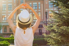 La fille dans le chapeau montre le coeur avec ses doigts, se tient avec elle de retour, regarde le coucher du soleil, style de vi photographie stock La fille dans le chapeau montre le coeur avec ses doigts, se tient avec elle de retour, regarde le coucher du soleil, style de vi photographie stock