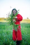 La fille dans la robe rouge fait une guirlande de l'herbe et des fleurs sauvages sur le fond d'un pré vert photos libres de droits La fille dans la robe rouge fait une guirlande de l'herbe et des fleurs sauvages sur le fond d'un pré vert photos libres de droits