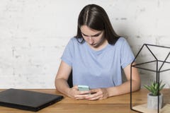 La fille s'assied à la table et tient le téléphone dans des ses mains Elle écrit des messages Il y a un ordinateur portable sur l images stock La fille s'assied à la table et tient le téléphone dans des ses mains Elle écrit des messages Il y a un ordinateur portable sur l images stock