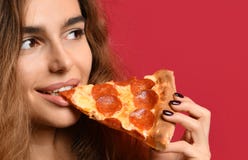 La giovane bella donna mangia la fetta di pizza di merguez con gli occhi chiusi che sorride sui rossi carmini fotografie stock libere da diritti La giovane bella donna mangia la fetta di pizza di merguez con gli occhi chiusi che sorride sui rossi carmini fotografie stock libere da diritti