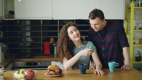 La giovane donna caucasica felice riccia attraente sta sedendosi nella cucina che manda un sms a qualcuno tramite smartphone, il stock footage La giovane donna caucasica felice riccia attraente sta sedendosi nella cucina che manda un sms a qualcuno tramite smartphone, il stock footage