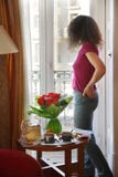 La giovane donna sta ed esamina fuori la finestra la tavola immagine stock libera da diritti La giovane donna sta ed esamina fuori la finestra la tavola immagine stock libera da diritti