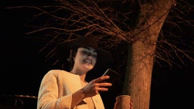 La giovane ragazza castana alla notte in un cappello esamina il telefono e beve il caffè sotto l'albero video d archivio La giovane ragazza castana alla notte in un cappello esamina il telefono e beve il caffè sotto l'albero video d archivio