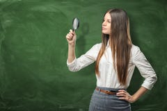 La jeune femme dans l'habillement officiel regarde avec une loupe sur le fond du tableau noir vert Image stock La jeune femme dans l'habillement officiel regarde avec une loupe sur le fond du tableau noir vert Image stock
