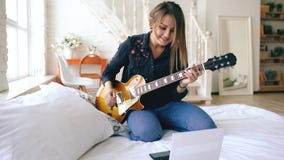 La jeune fille attirante apprenant à jouer la guitare électrique avec le carnet s'asseyent sur le lit dans la chambre à coucher à photo libre de droits La jeune fille attirante apprenant à jouer la guitare électrique avec le carnet s'asseyent sur le lit dans la chambre à coucher à photo libre de droits