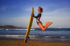 La jeune fille heureuse et attirante de surfer sautant haut dans le ciel tenant le panneau de ressac avant de surfer à la belle p image stock La jeune fille heureuse et attirante de surfer sautant haut dans le ciel tenant le panneau de ressac avant de surfer à la belle p image stock