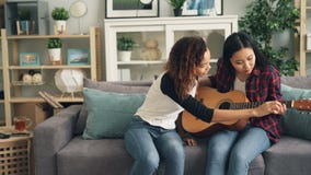 La muchacha afroamericana alegre está enseñando a su amigo asiático a tocar la guitarra en casa Las mujeres jovenes se están sent almacen de video La muchacha afroamericana alegre está enseñando a su amigo asiático a tocar la guitarra en casa Las mujeres jovenes se están sent almacen de video