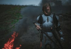 La muchacha en la imagen del arco del ` de Jeanne d en armadura y con la espada en sus manos se opone al fondo del fuego y del hu fotos de archivo La muchacha en la imagen del arco del ` de Jeanne d en armadura y con la espada en sus manos se opone al fondo del fuego y del hu fotos de archivo