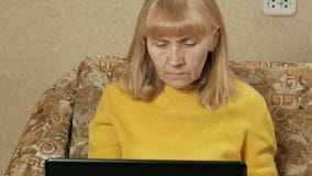 La mujer envejecida trabaja cuidadosamente en un ordenador portátil en casa en el sofá Ella está mecanografiando en el teclado y metrajes La mujer envejecida trabaja cuidadosamente en un ordenador portátil en casa en el sofá Ella está mecanografiando en el teclado y metrajes