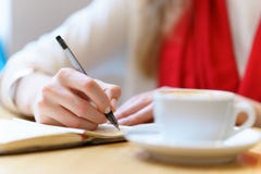 La mujer europea con la bufanda roja está escribiendo por la pluma algo en la libreta cerca de la taza de café blanca en la tabla Foto de archivo La mujer europea con la bufanda roja está escribiendo por la pluma algo en la libreta cerca de la taza de café blanca en la tabla Foto de archivo