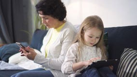 La mujer mayor se está sentando en el sofá y está mirando smartphone mientras que la chica joven está mirando algo interesante en metrajes La mujer mayor se está sentando en el sofá y está mirando smartphone mientras que la chica joven está mirando algo interesante en metrajes