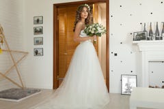 La novia en un vestido de boda hermoso con un pelo rizado largo se coloca en el dormitorio y mira la cámara foto de archivo libre de regalías La novia en un vestido de boda hermoso con un pelo rizado largo se coloca en el dormitorio y mira la cámara foto de archivo libre de regalías