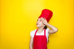 La petite fille dans la fille du costume d'un chef rouge est fatiguée de la cuisson, essuie son front de main sur le fond jaune a photo libre de droits La petite fille dans la fille du costume d'un chef rouge est fatiguée de la cuisson, essuie son front de main sur le fond jaune a photo libre de droits