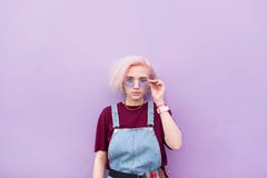 La ragazza alla moda con capelli e gli occhiali da sole rosa è un fondo porpora e sguardi alla macchina fotografica fotografie stock La ragazza alla moda con capelli e gli occhiali da sole rosa è un fondo porpora e sguardi alla macchina fotografica fotografie stock