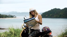 La ragazza bionda in un cappello, si siede su una bici ed esamina il telefono e una mappa, esamina l'itinerario in Asia video d archivio La ragazza bionda in un cappello, si siede su una bici ed esamina il telefono e una mappa, esamina l'itinerario in Asia video d archivio