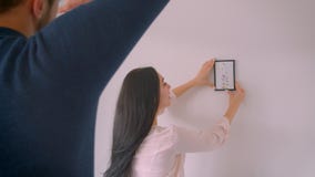 La ragazza caucasica sta scegliendo il posto per l'immagine incorniciata sulla parete bianca mentre il suo ragazzo fa la forma de La ragazza caucasica sta scegliendo il posto per l'immagine incorniciata sulla parete bianca mentre il suo ragazzo fa la forma de