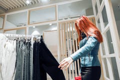 La ragazza rossa dei capelli in un bomber blu sceglie i pantaloni da vendere fotografia stock La ragazza rossa dei capelli in un bomber blu sceglie i pantaloni da vendere fotografia stock