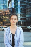 La ragazza teenager sveglia in rivestimento sta sulla via contro un grattacielo moderno immagine stock libera da diritti La ragazza teenager sveglia in rivestimento sta sulla via contro un grattacielo moderno immagine stock libera da diritti