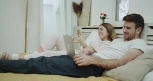 La rutina de la mañana para un par en cama, la mujer que lee un libro y su socio están mirando en el ordenador portátil Tirado en almacen de metraje de vídeo La rutina de la mañana para un par en cama, la mujer que lee un libro y su socio están mirando en el ordenador portátil Tirado en almacen de metraje de vídeo