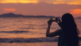 La siluetta di giovane donna turistica fotografa la vista di oceano con lo smartphone durante il tramonto alla spiaggia fotografie stock La siluetta di giovane donna turistica fotografa la vista di oceano con lo smartphone durante il tramonto alla spiaggia fotografie stock