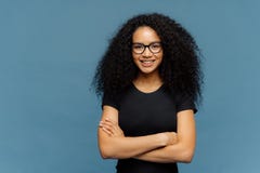 La taille a tiré de la femme afro-américaine de sourire fait plier des bras, porte des lunettes et le T-shirt noir occasionnel, a image stock La taille a tiré de la femme afro-américaine de sourire fait plier des bras, porte des lunettes et le T-shirt noir occasionnel, a image stock