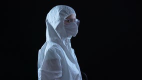 Lab worker in protective suit and mask turning to camera against dark background
