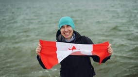 Langzaam motieportret van de gelukkige jonge mens die grote textielvlag van Canada houden, camera bekijken en met water glimlache stock video Langzaam motieportret van de gelukkige jonge mens die grote textielvlag van Canada houden, camera bekijken en met water glimlache stock video