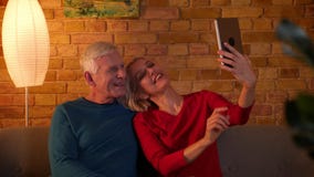 Lanzamiento del primer de los pares felices mayores que toman selfies en la tableta que se sienta en el sofá dentro en un apartam almacen de video Lanzamiento del primer de los pares felices mayores que toman selfies en la tableta que se sienta en el sofá dentro en un apartam almacen de video