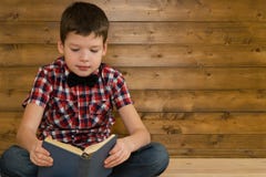 Le garçon s'assied sur le plancher lisant un livre sur le fond d'un mur en bois Image libre de droits Le garçon s'assied sur le plancher lisant un livre sur le fond d'un mur en bois Image libre de droits