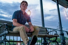 Le jeune homme repose heureusement sur le banc de parc avec s'allumer de poteau léger de lancement le ciel de soirée derrière lui photos libres de droits Le jeune homme repose heureusement sur le banc de parc avec s'allumer de poteau léger de lancement le ciel de soirée derrière lui photos libres de droits