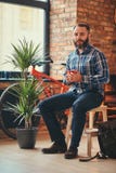 Le mâle barbu beau de hippie dans une chemise bleue et des jeans d'ouatine tient une tasse de café de matin tout en se reposant s photos stock Le mâle barbu beau de hippie dans une chemise bleue et des jeans d'ouatine tient une tasse de café de matin tout en se reposant s photos stock