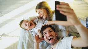 Le sourire parents avec le bébé prenant la photo de famille de selfie sur le lit à la maison Photos libres de droits Le sourire parents avec le bébé prenant la photo de famille de selfie sur le lit à la maison Photos libres de droits