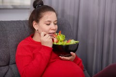 Le tir de la femme mignonne enceinte de brune attrayante a plaisir à manger de la salade fraîche des tomates et de la laitue, pos photographie stock Le tir de la femme mignonne enceinte de brune attrayante a plaisir à manger de la salade fraîche des tomates et de la laitue, pos photographie stock