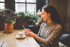 Les belles utilisations de jeune fille, types textotent à un téléphone portable à une table en bois près de la fenêtre et boivent images stock Les belles utilisations de jeune fille, types textotent à un téléphone portable à une table en bois près de la fenêtre et boivent images stock
