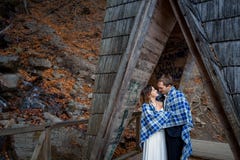 Les jeunes mariés heureux enveloppés dans la couverture étreignent doucement sur le pont en bois aux montagnes Fond de forêt d'au Images stock Les jeunes mariés heureux enveloppés dans la couverture étreignent doucement sur le pont en bois aux montagnes Fond de forêt d'au Images stock