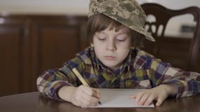 Leuke grappig weinig jongen in geruit overhemd en militaire hoed van zijn vader die een brievenzitting schrijven thuis bij de lij stock footage Leuke grappig weinig jongen in geruit overhemd en militaire hoed van zijn vader die een brievenzitting schrijven thuis bij de lij stock footage