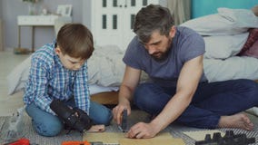Little boy is learning to use electric screwdriver while his dad is explaining how to work with gun and fix