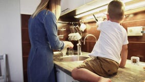 Little boy son sits on table and helps mom in the kitchen sink