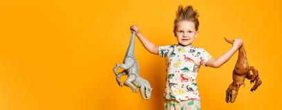 Little cheerful boy holds a plastic dinosaur in each hand. The boy is dressed in a suit with pictures of dinosaurs.
