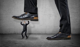 Little frightened worker under feet of huge businessman on grey wall background stock image Little frightened worker under feet of huge businessman on grey wall background stock image