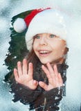 Little girl in santa hat is looking through snowy window