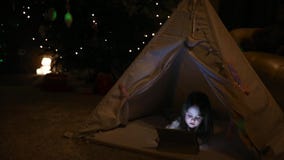 Little girl sitting at home in a tent against the background of a Christmas tree playing games on an electronic tablet stock video Little girl sitting at home in a tent against the background of a Christmas tree playing games on an electronic tablet stock video