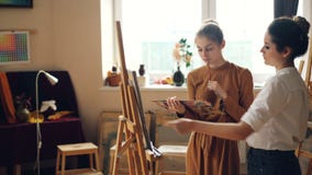 Lo studente di arte allegro sta lavorando alla pittura dell'immagine con la spazzola su tela mentre il suo insegnante sta control stock footage Lo studente di arte allegro sta lavorando alla pittura dell'immagine con la spazzola su tela mentre il suo insegnante sta control stock footage
