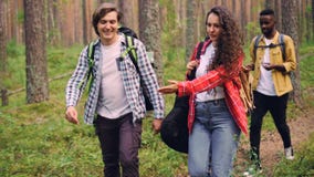 Los amigos sonrientes de la gente joven están caminando en bosque con las mochilas, la muchacha atractiva está llevando la guitar metrajes Los amigos sonrientes de la gente joven están caminando en bosque con las mochilas, la muchacha atractiva está llevando la guitar metrajes